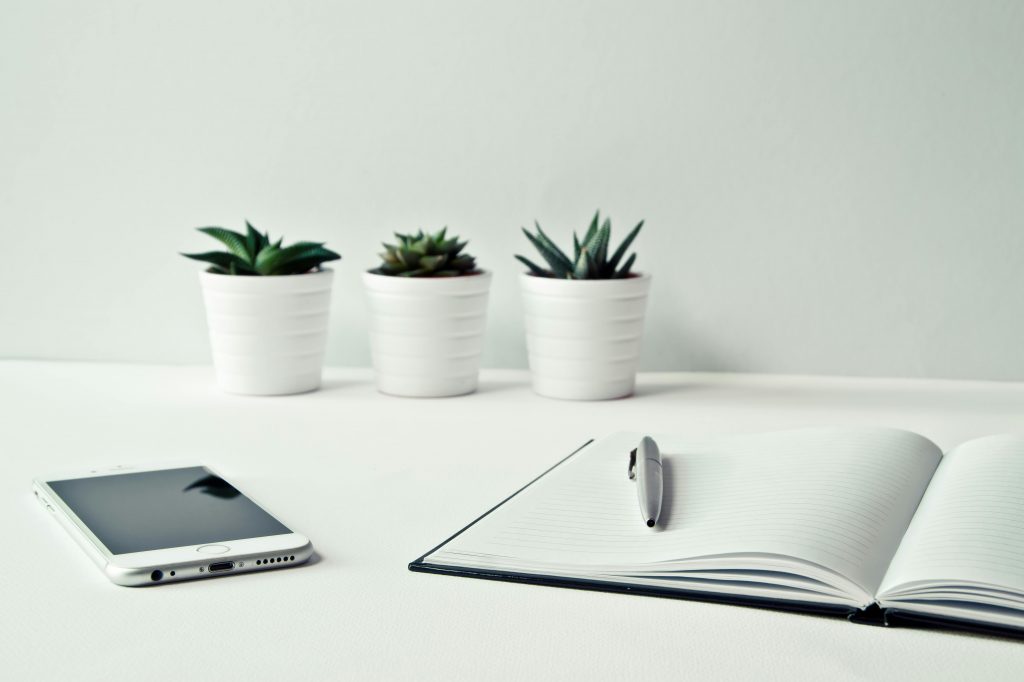 A clean, minimalist workspace featuring a smartphone, open notepad, pen, and potted succulents.